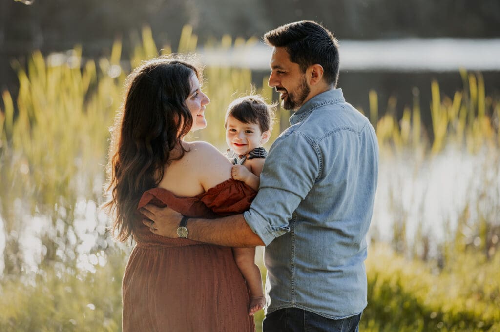 Family photography at Tree Tops Park in Davie Florida, parents holding baby by the lake with golden light and tall grass in the background