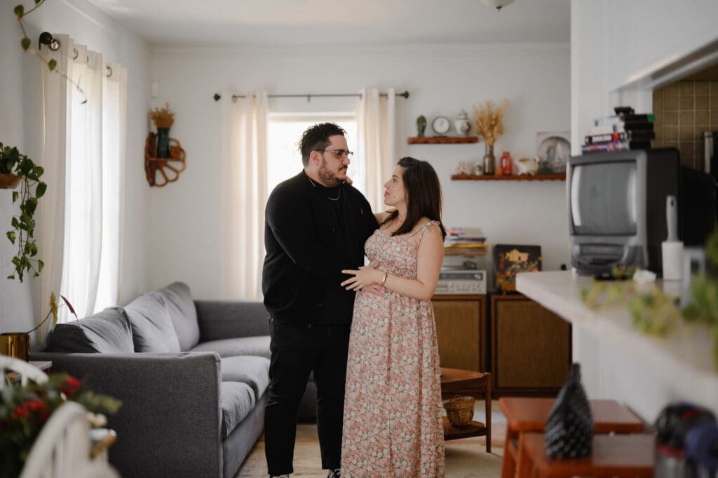 In home maternity session with an expecting couple standing together in their living room, photographed indoors in natural light

