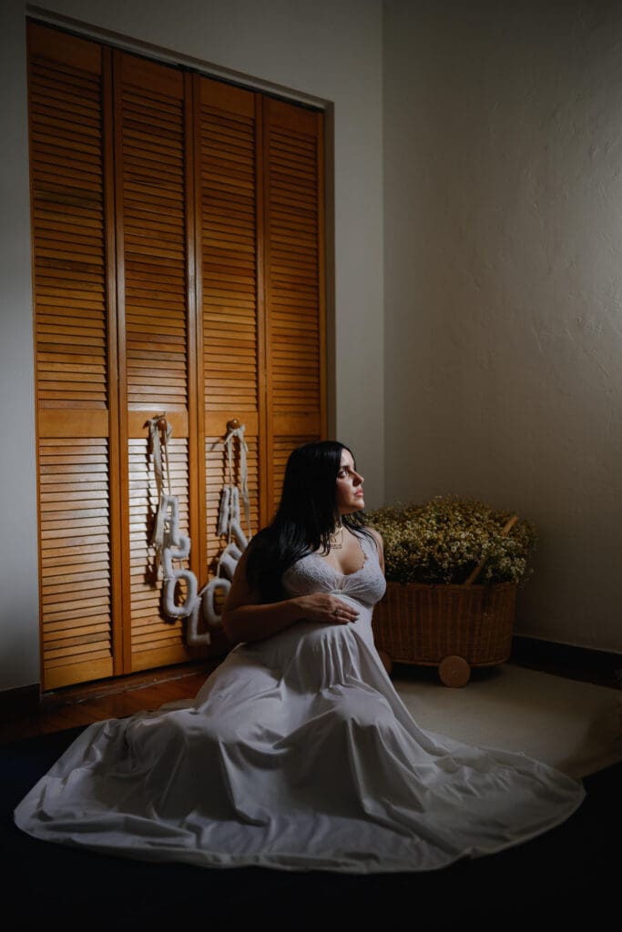 Black and white maternity portrait of a pregnant woman wearing a flowing dress and sitting on the floor by a window