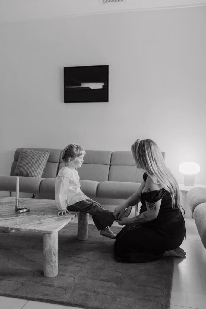 Black and white lifestyle family photo of a mother kneeling to help her child indoors in a minimal living room