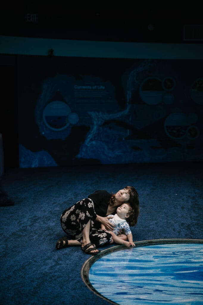 Lifestyle family photo of a mother and young child sitting together indoors, wearing comfortable everyday clothing in a dark blue space