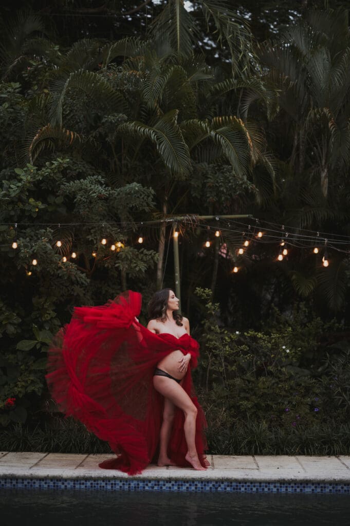 Lifestyle maternity photography showing a pregnant woman in a flowing red dress with movement near a pool and tropical plants