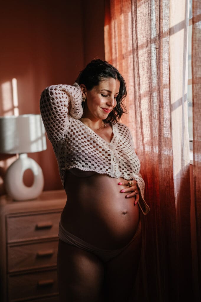 In home maternity session featuring a pregnant woman standing by a window in soft natural light, photographed indoors