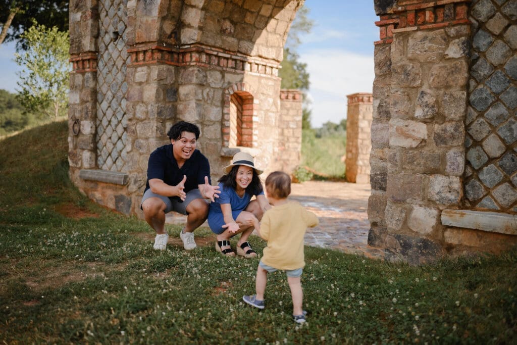 Parents encouraging their toddler to run toward them during an outdoor family photo session at BridgeWay Station in Greenville, South Carolina.